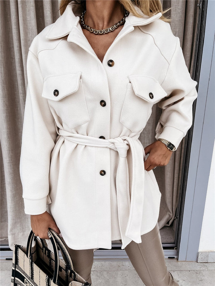 Frau in cremefarbener Jacke mit Gürtel, Knöpfen und Taschen, trägt eine Tasche und eine Uhr.