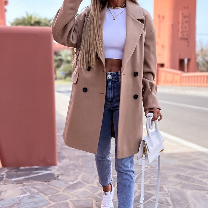 Frau in beigem Mantel, weißem Top, Jeans und weißen Sneakers, hält weiße Handtasche.