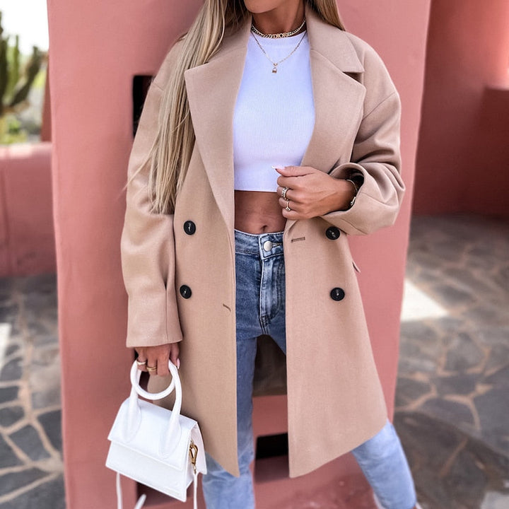 Frau in beigem Wollmantel, weißem Crop-Top, Jeans, hält weiße Handtasche, modisch, elegant.