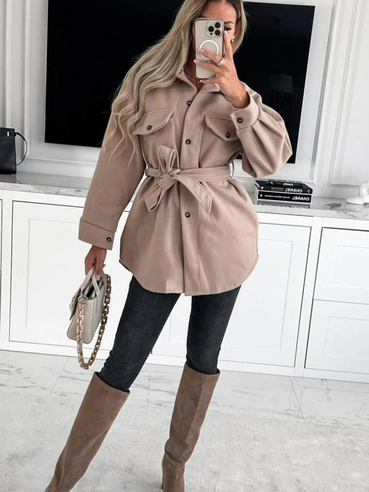 Frau in beiger Jacke mit Gürtel, schwarze Jeans, beige Stiefel, Handy, modisches Outfit.