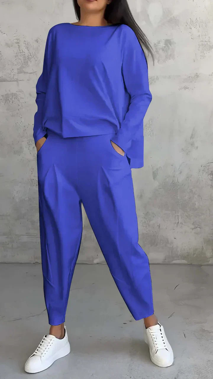 Frau in blauem Oversize-Anzug mit weißen Sneakers vor grauer Wand.