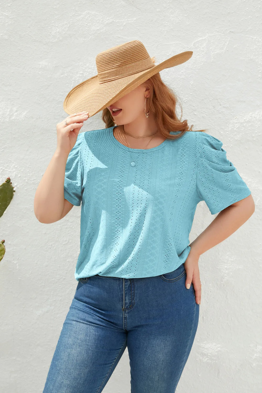 Frau mit Strohhut, blauer Spitzenbluse und Jeans vor weißer Wand.