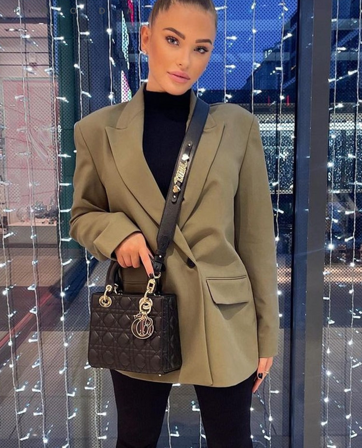 Frau mit olivgrünem Blazer und schwarzer Designer-Handtasche vor beleuchtetem Hintergrund.