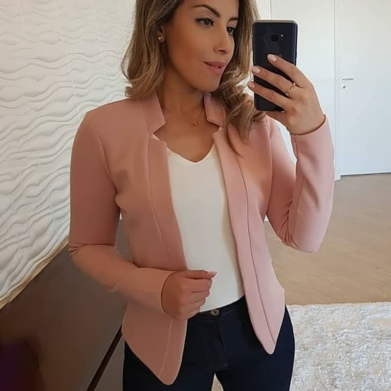 Frau in rosa Blazer, weißem Top, dunkler Jeans, Smartphone in der Hand, modernes Interieur.