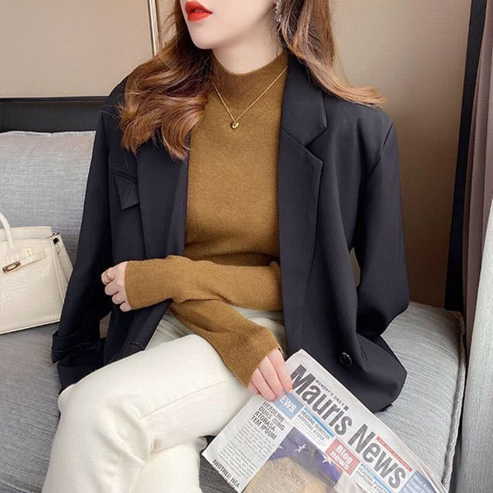 Frau in schwarzem Blazer, braunem Pullover, hält Zeitung, sitzt auf Sofa, modisch, elegant.