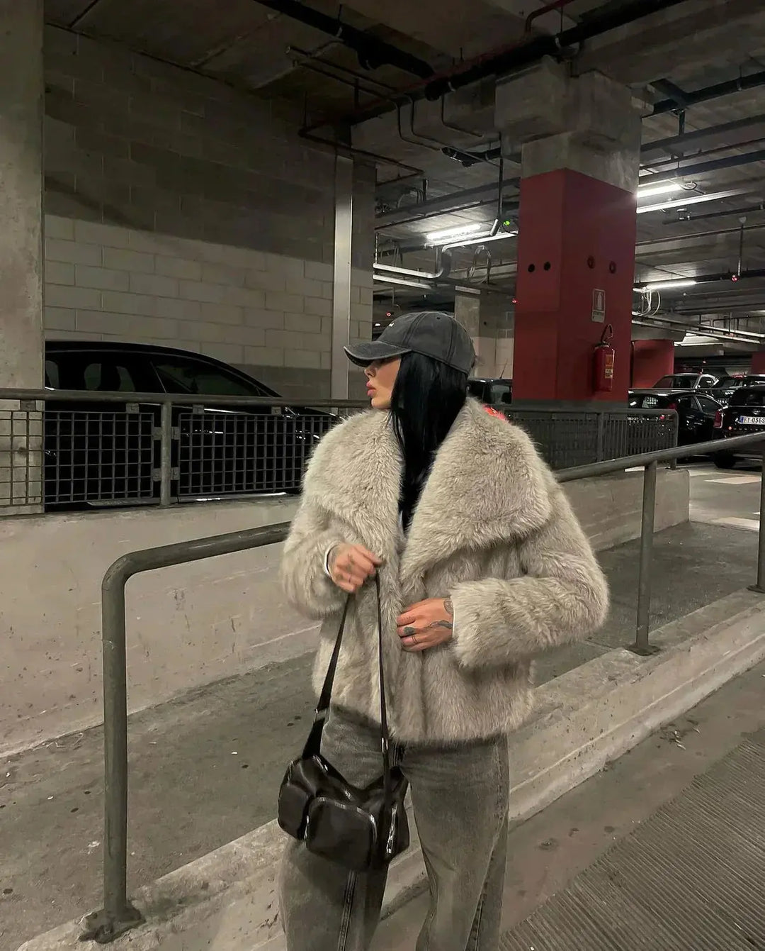 Frau in grauem Pelzmantel und Jeans mit schwarzer Tasche in Tiefgarage.