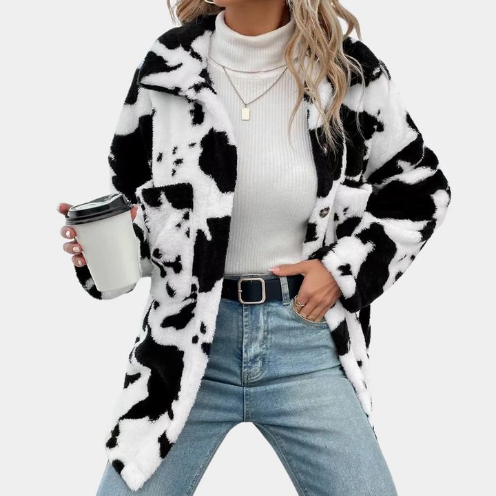Frau in schwarz-weißer Kuhfelljacke, weißem Rollkragenpullover, Jeans, hält Kaffeebecher.