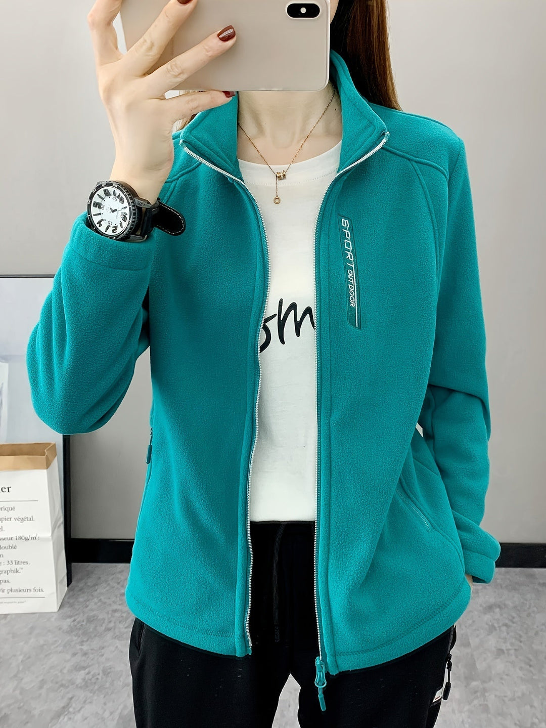 Frau in türkisfarbener Fleecejacke, weißes T-Shirt, schwarze Hose, Armbanduhr, Smartphone.