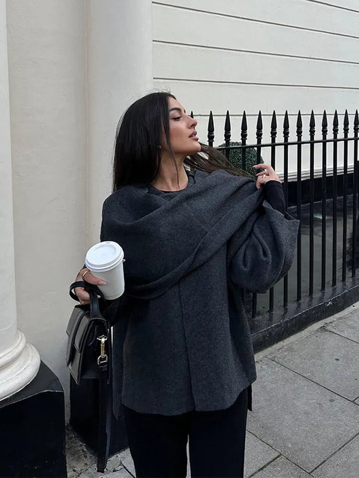 Frau in grauem Wollmantel mit Kaffeebecher und schwarzer Tasche vor Zaun und Gebäude.