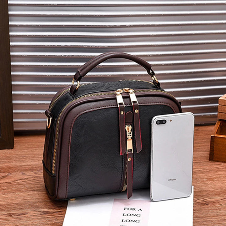 Schwarze Lederhandtasche mit goldenen Reißverschlüssen neben einem Smartphone auf Holztisch.