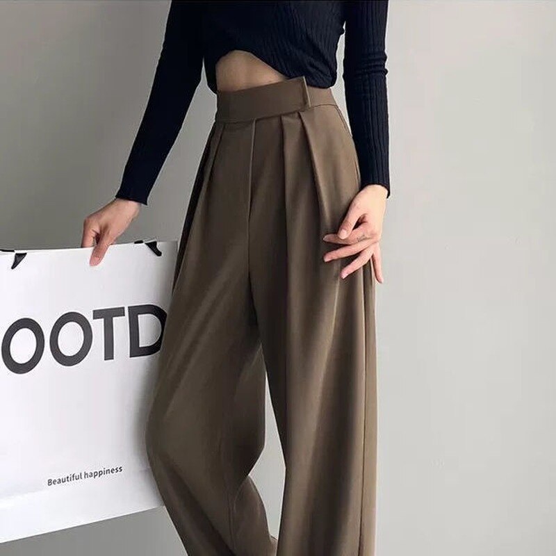 Frau in brauner, weiter Hose und schwarzem Oberteil hält Schild, modisch, elegant, minimalistisch.