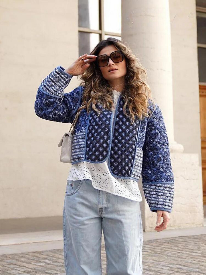 Frau in blauer Steppjacke, weißer Bluse und Jeans, trägt Sonnenbrille, steht draußen.