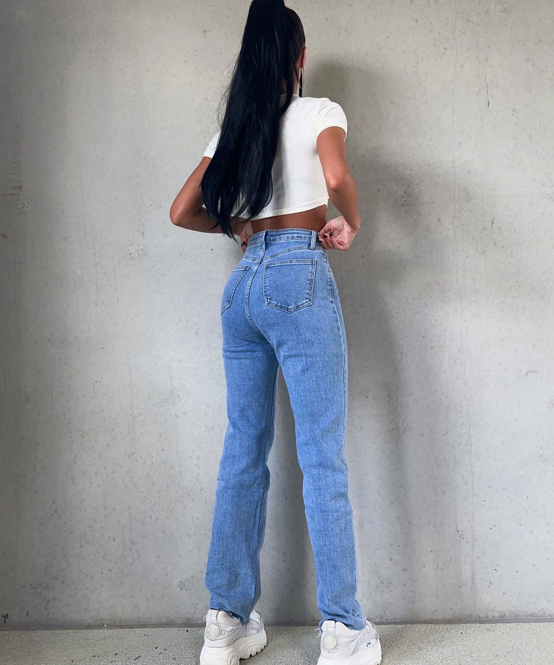 Frau in blauer Jeans und weißem Crop-Top, Rückansicht, vor grauer Wand.