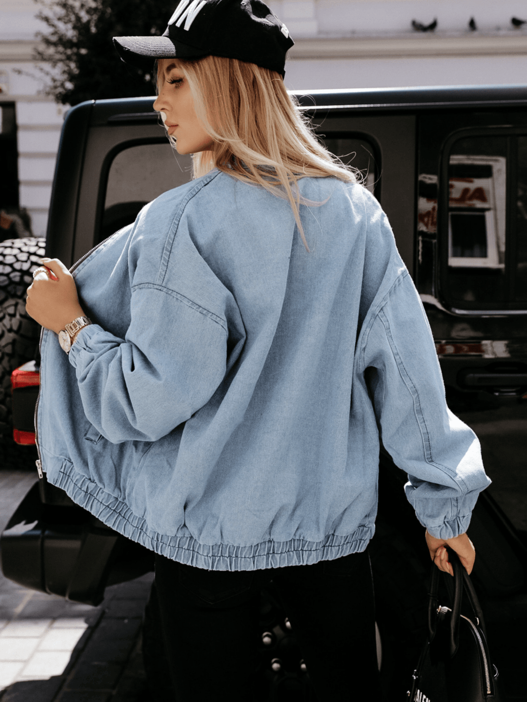 Frau in blauer Jeansjacke und schwarzer Kappe vor schwarzem SUV, modisch, urbaner Stil.