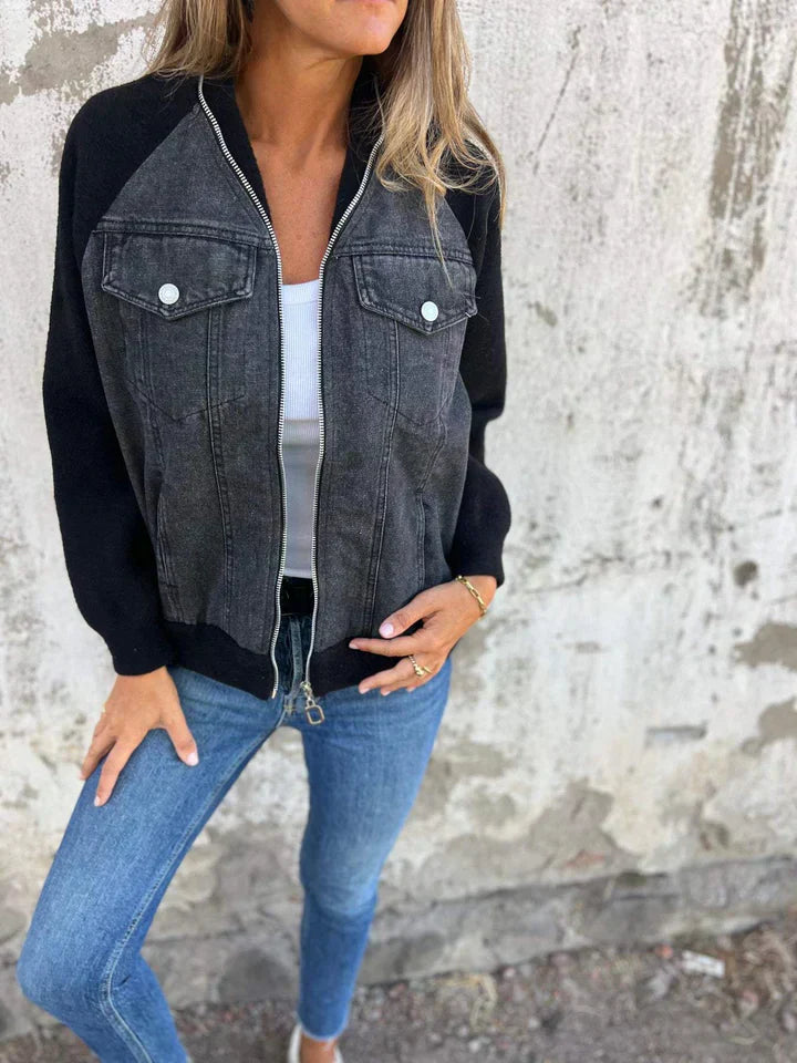 Frau in schwarzer Jeansjacke, weißem Top und blauer Jeans vor Betonwand.
