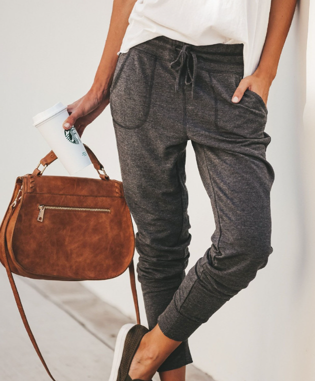 Frau in grauer Jogginghose mit brauner Ledertasche und Kaffeebecher, lässiger Streetstyle.