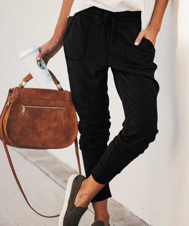Frau in schwarzer Jogginghose, brauner Umhängetasche, hält Kaffeebecher, lässiger Stil.