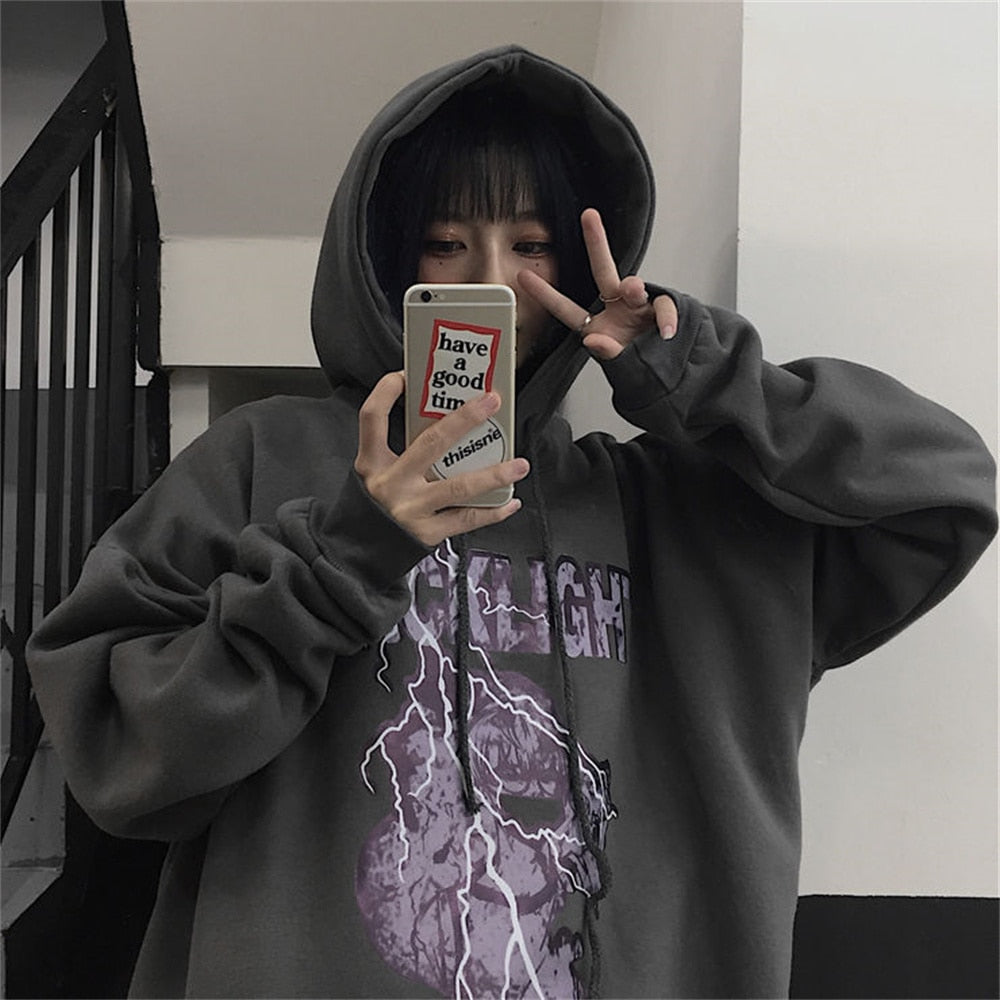 Frau in grauem Kapuzenpullover mit Blitzmotiv, Selfie, Smartphone, Treppe im Hintergrund.
