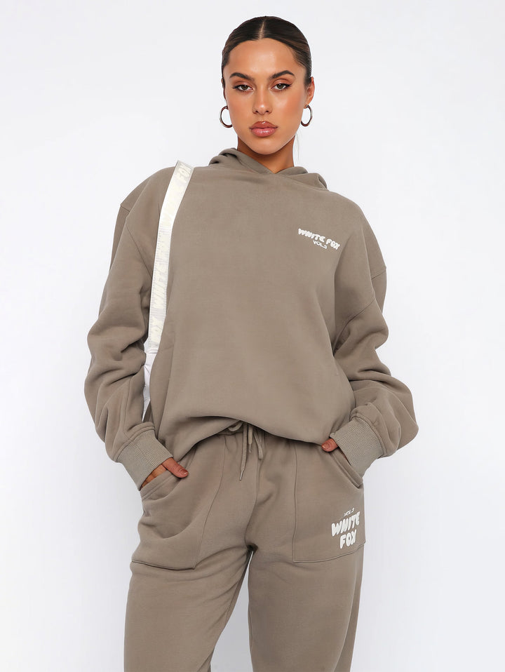 Frau in beigem Hoodie und Jogginghose, White Fox Logo, lässige Mode, Streetwear-Outfit.