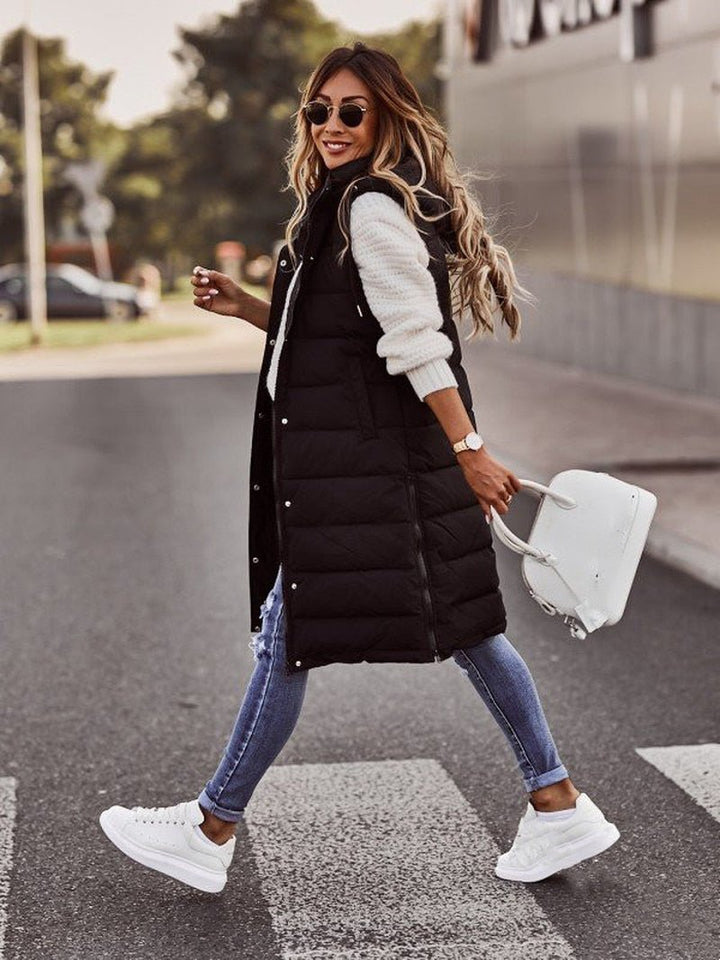 Frau in schwarzer, knielanger Steppweste, weiße Sneakers, Jeans, weiße Tasche, Straße.
