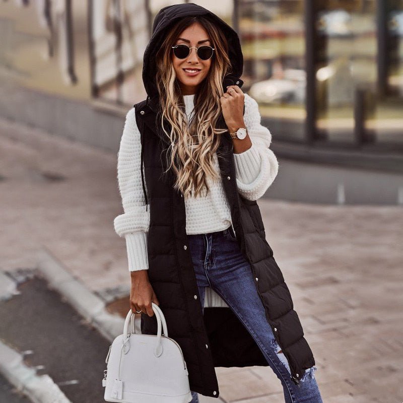 Frau in schwarzer, knielanger Steppweste mit Kapuze, weißem Pullover, Jeans und Sonnenbrille.