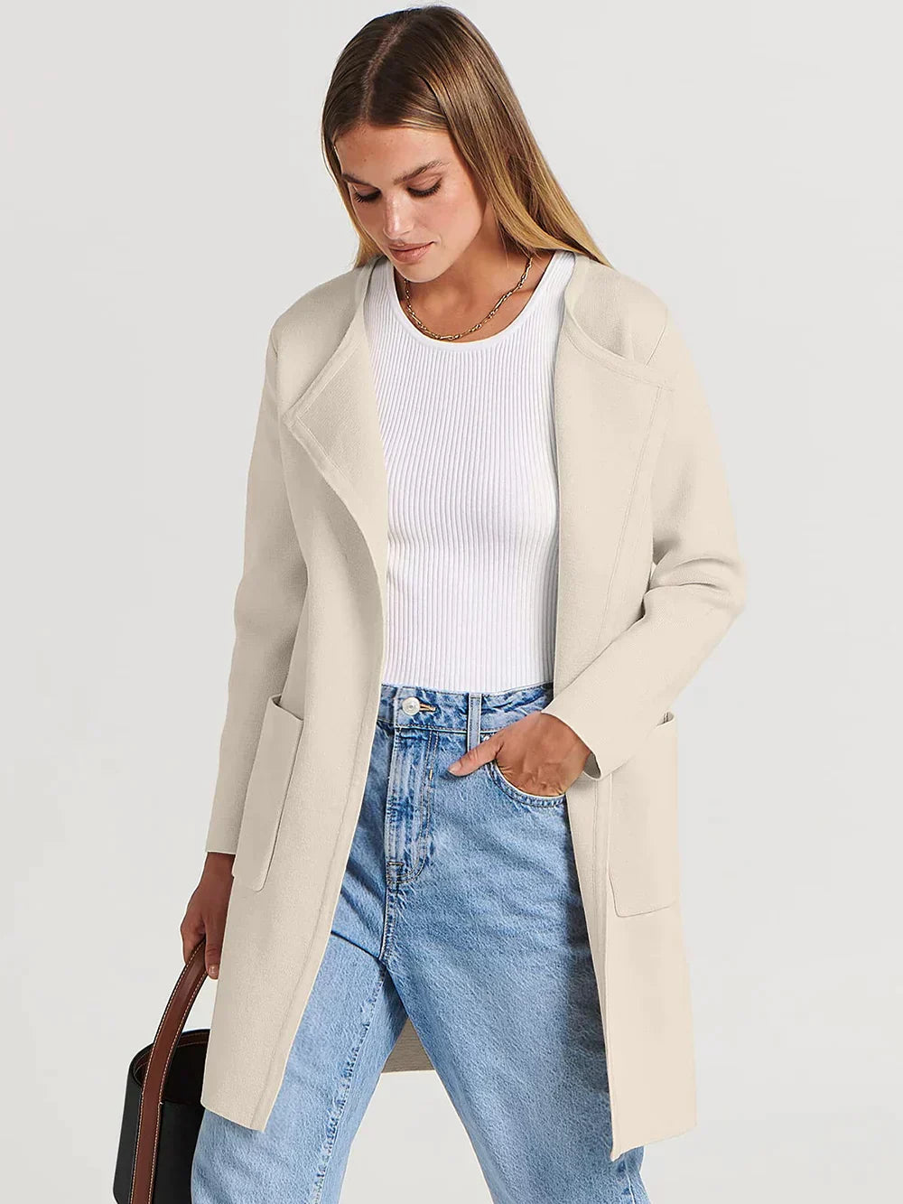 Frau in cremefarbenem Mantel, weißem Top und blauer Jeans, hält schwarze Tasche.