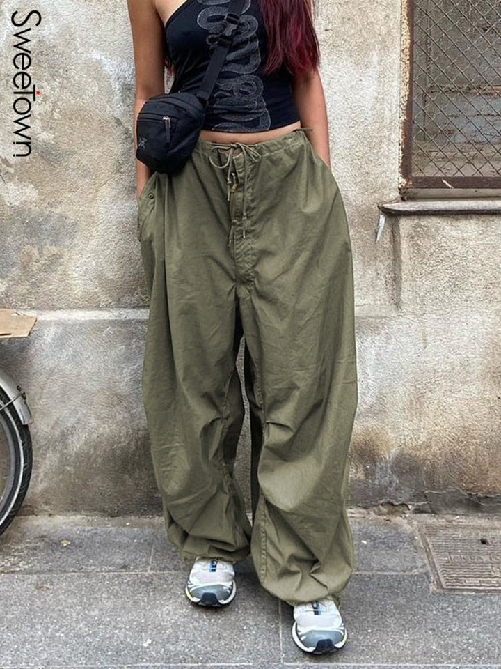 Frau in olivgrüner Baggy-Hose, schwarzem Top und Sneakern vor Betonwand.
