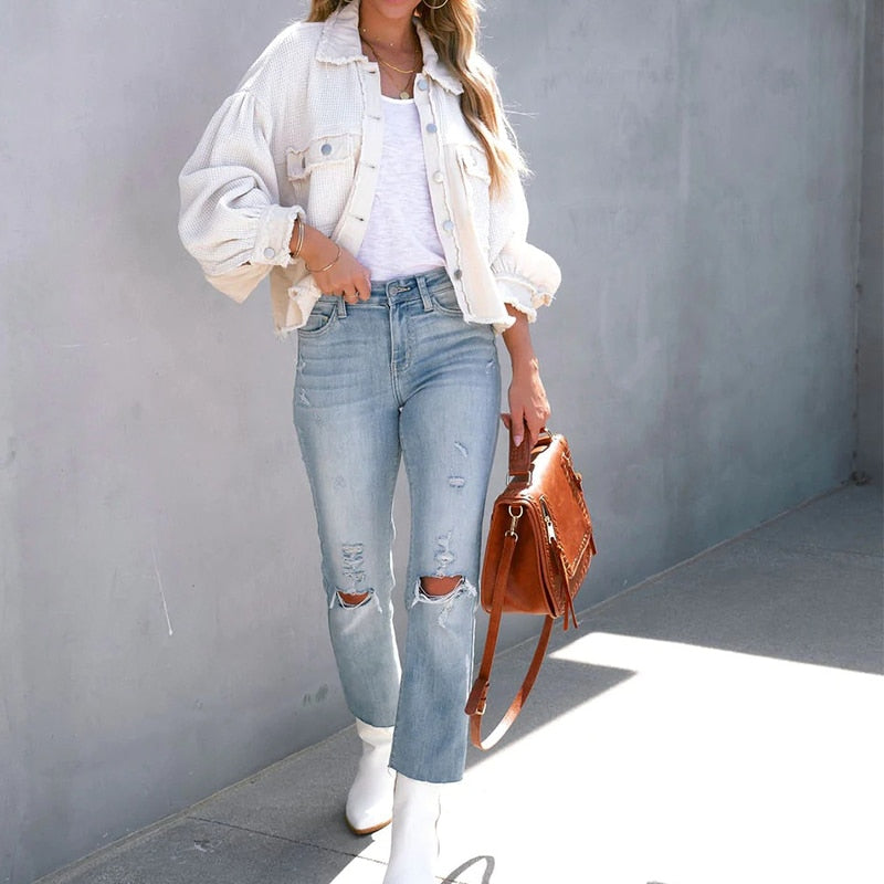 Frau in weißer Jacke, zerrissener Jeans, brauner Tasche, weißen Stiefeln vor grauer Wand.