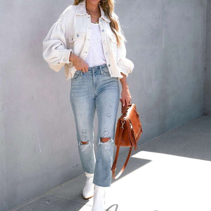 Frau in weißer Jacke, zerrissener Jeans, brauner Tasche, weißen Stiefeln vor grauer Wand.