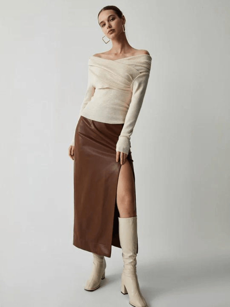 Frau in cremefarbenem Pullover, braunem Lederrock mit Schlitz und beigen Stiefeln.