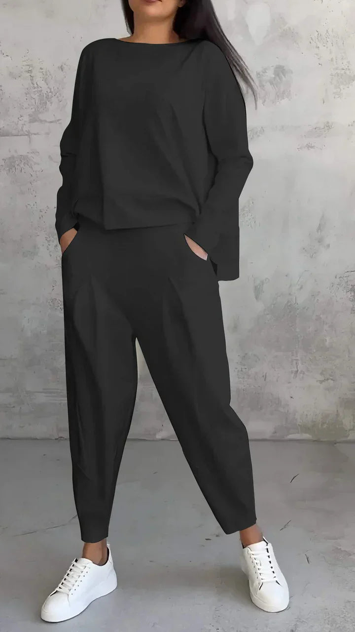 Frau in schwarzem Oversize-Zweiteiler mit weißen Sneakers vor grauer Wand.