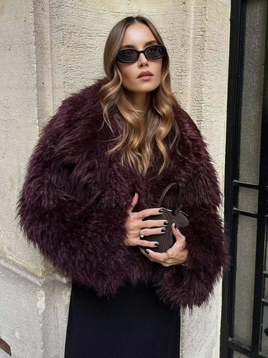 Frau in dunkelrotem Kunstpelzmantel, schwarze Sonnenbrille, hält schwarze Handtasche.