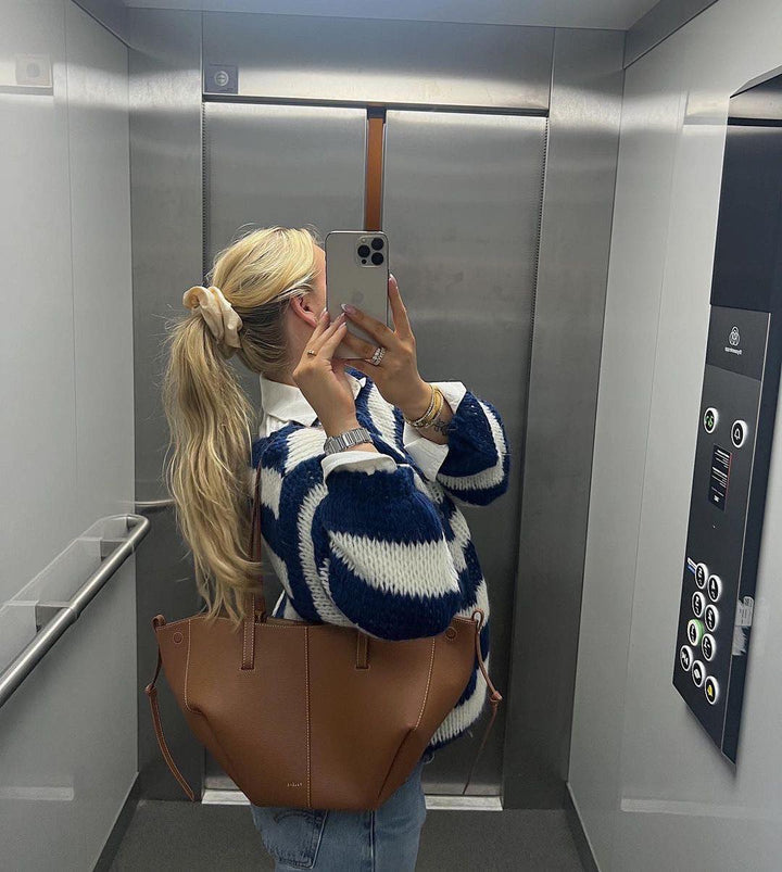 Frau im Aufzug mit blau-weißem Pullover, brauner Ledertasche, iPhone, blonden Haaren.