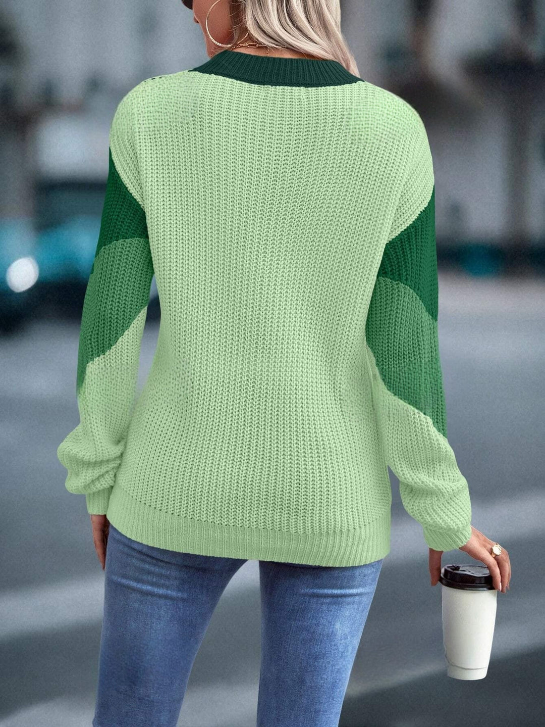 Frau in grünem Strickpullover und Jeans hält Kaffeebecher, modisch, urbaner Hintergrund.