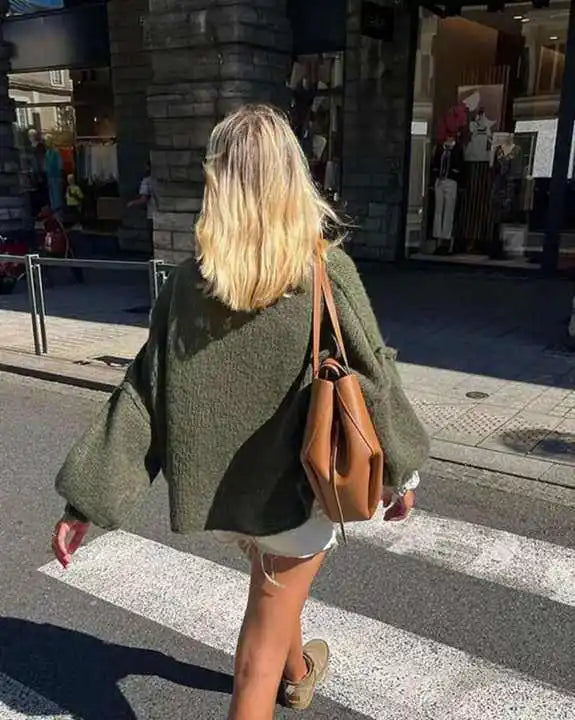 Frau mit grünem Strickpullover und brauner Ledertasche überquert Straße bei Sonnenschein.