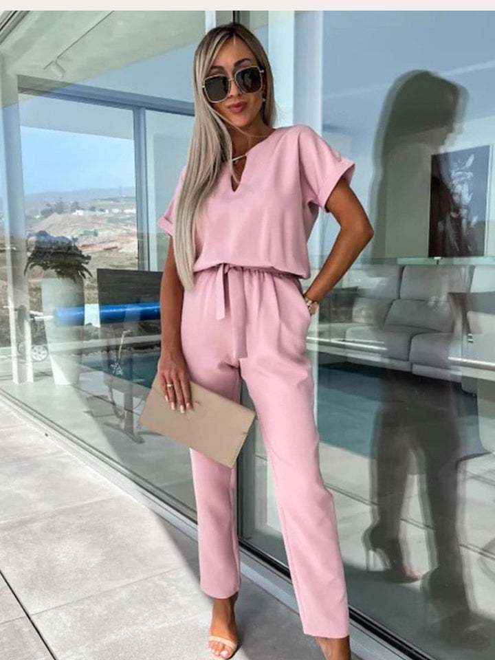 Frau in rosa Jumpsuit mit Sonnenbrille, beige Clutch, moderne Terrasse im Hintergrund.