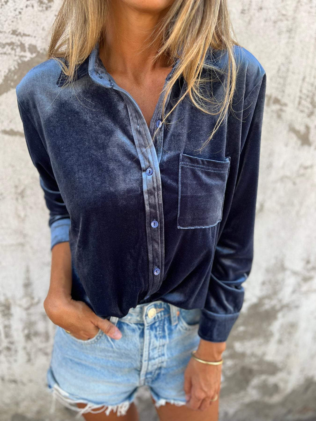 Frau in blauer Samtbluse und Jeansshorts vor Betonwand.