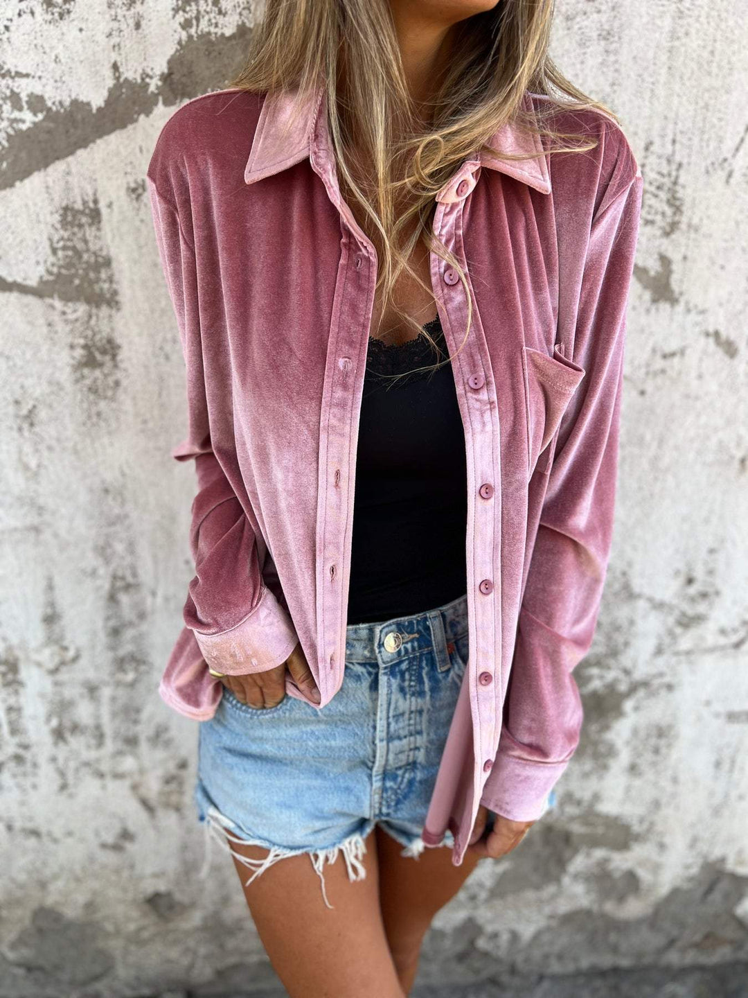 Frau in rosa Samthemd und Jeansshorts vor Betonwand.