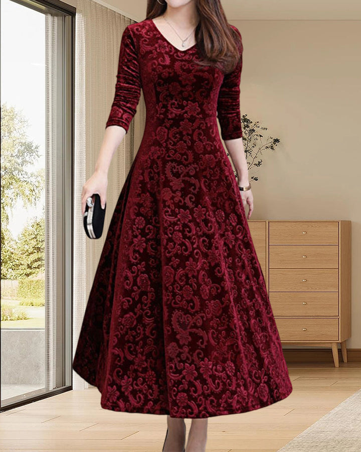 Frau in elegantem, rotem Samtkleid mit floralen Mustern, stehend in modernem Wohnzimmer.