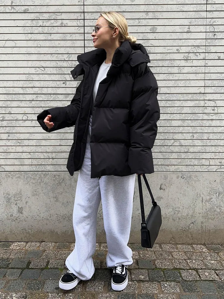 Frau in schwarzer Daunenjacke, grauer Jogginghose, Sneakers, vor gemauerter Wand.