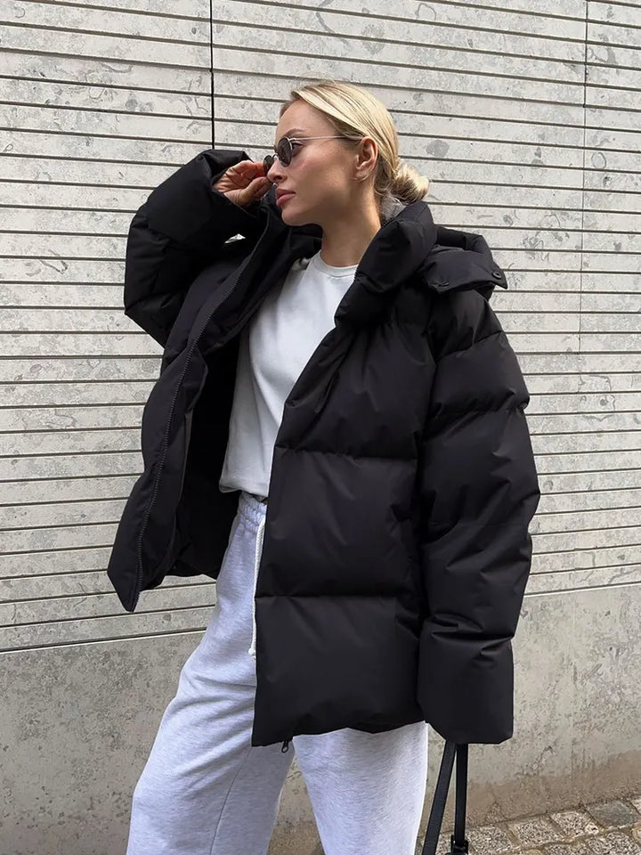 Frau in schwarzer Daunenjacke, weiße Jogginghose, Sonnenbrille, vor grauer Wand.