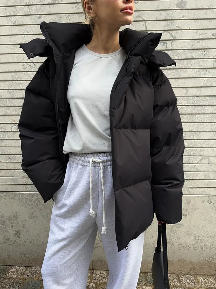 Frau in schwarzer Daunenjacke und weißem Sweatshirt vor Betonwand.