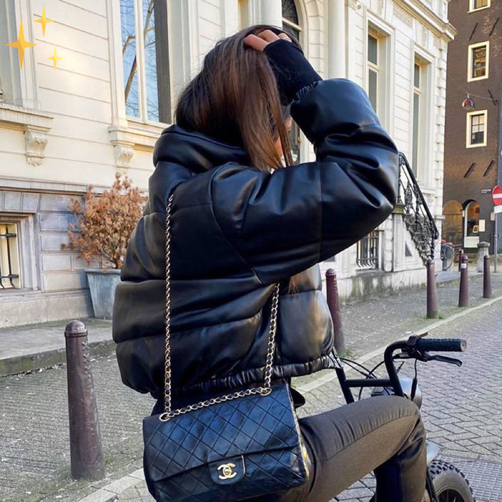 Frau in schwarzer Leder-Bomberjacke mit Chanel-Tasche auf Fahrrad in städtischer Umgebung.