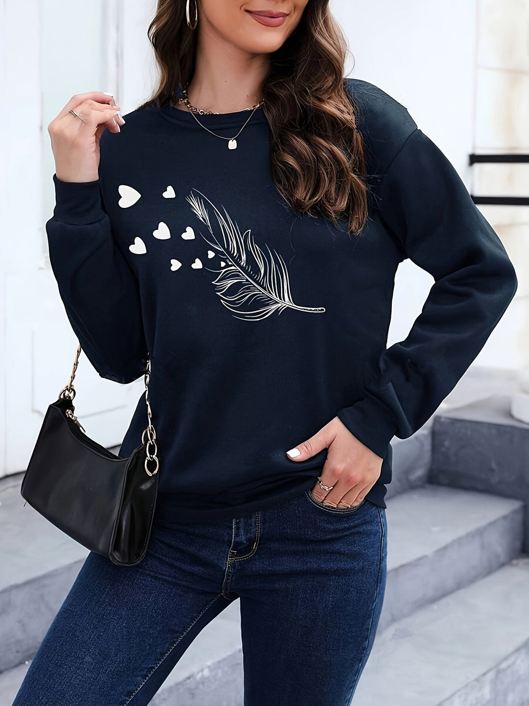 Frau in schwarzem Pullover mit Feder- und Herzmotiv, schwarze Handtasche, lässiger Stil.