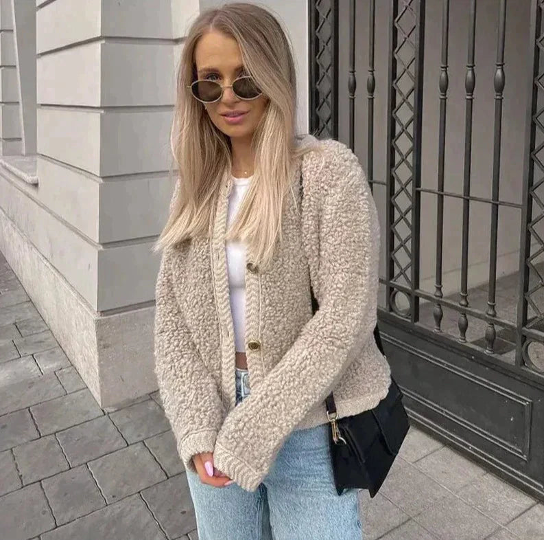 Frau in beiger Teddyjacke, weißem Top, blauer Jeans, Sonnenbrille, schwarze Tasche, draußen.