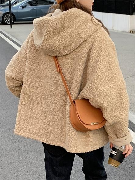 Frau in beiger Teddyjacke mit Kapuze, braune Umhängetasche, Kaffeebecher, Straße im Hintergrund.