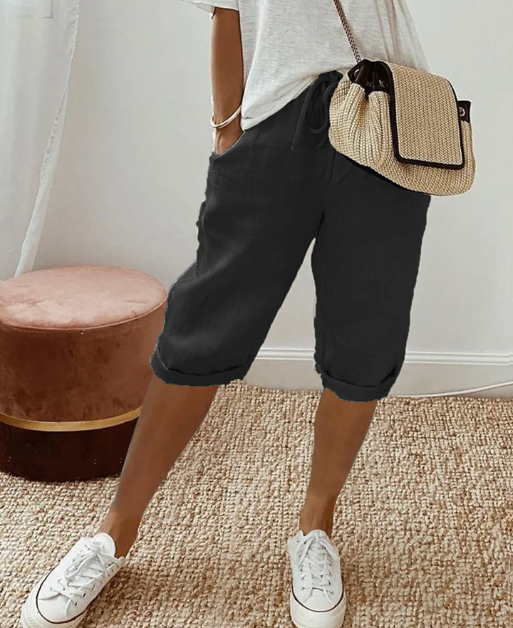 Frau in schwarzer Bermuda-Shorts, weißem T-Shirt, Strohtasche, weiße Sneaker, lässiger Stil.