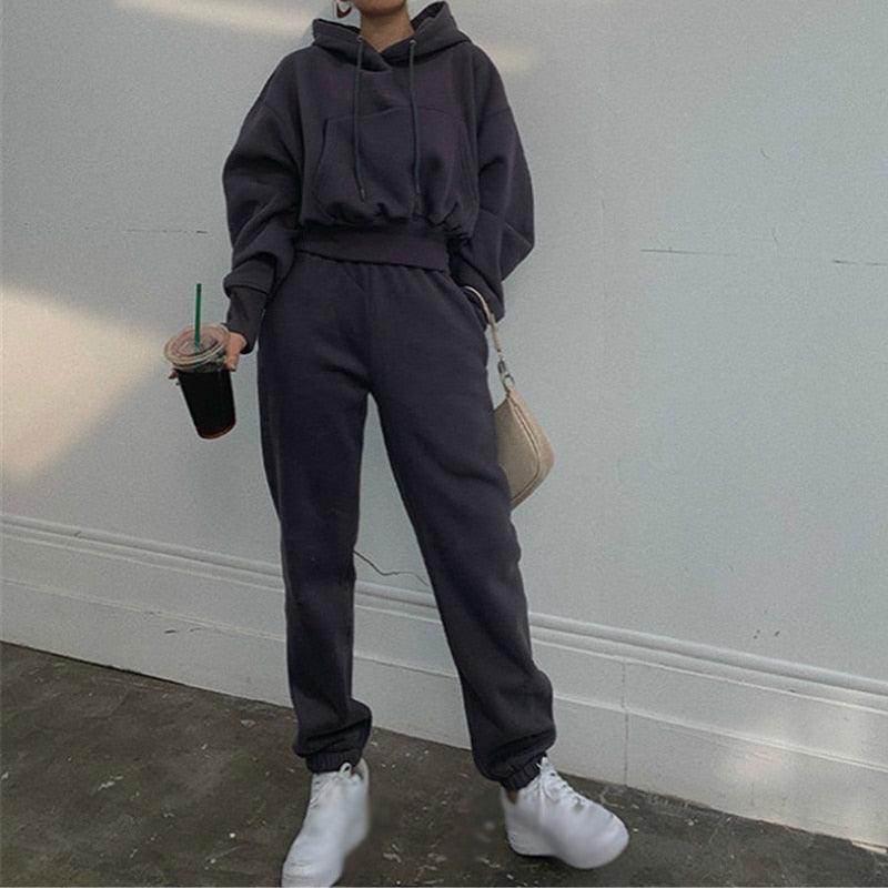 Frau in dunkelgrauem Hoodie und Jogginghose, hält Getränk, trägt weiße Sneaker.
