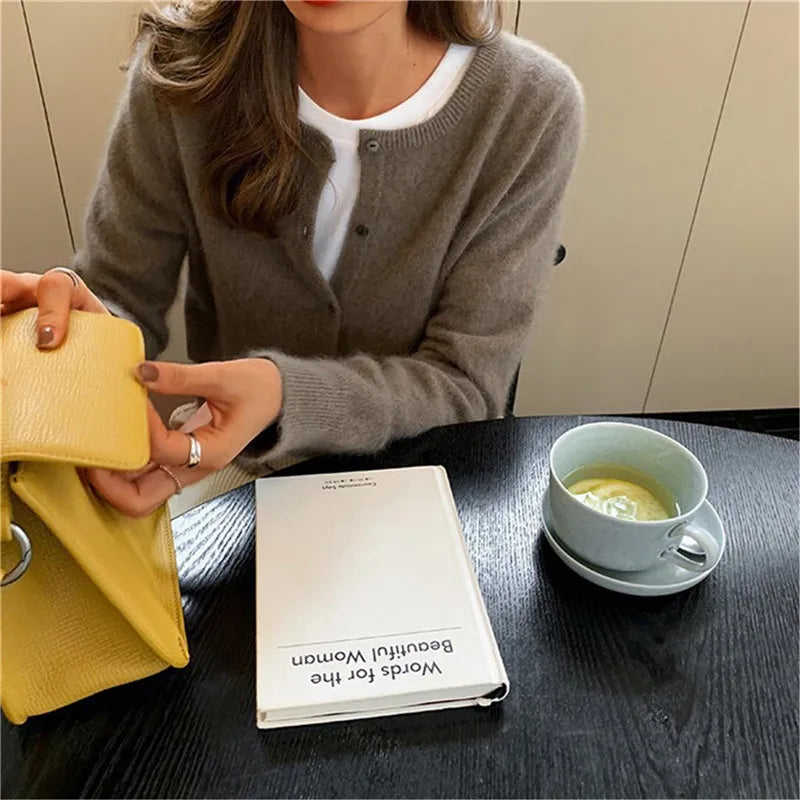 Frau mit grauem Strickcardigan, gelber Tasche, Buch und grünem Tee auf schwarzem Tisch.