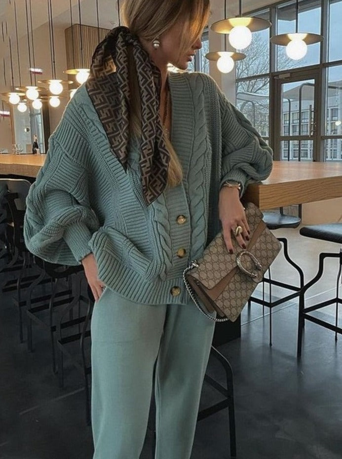 Frau in grünem Strickpullover und Hose, hält Designer-Handtasche, modernes Café-Interieur.